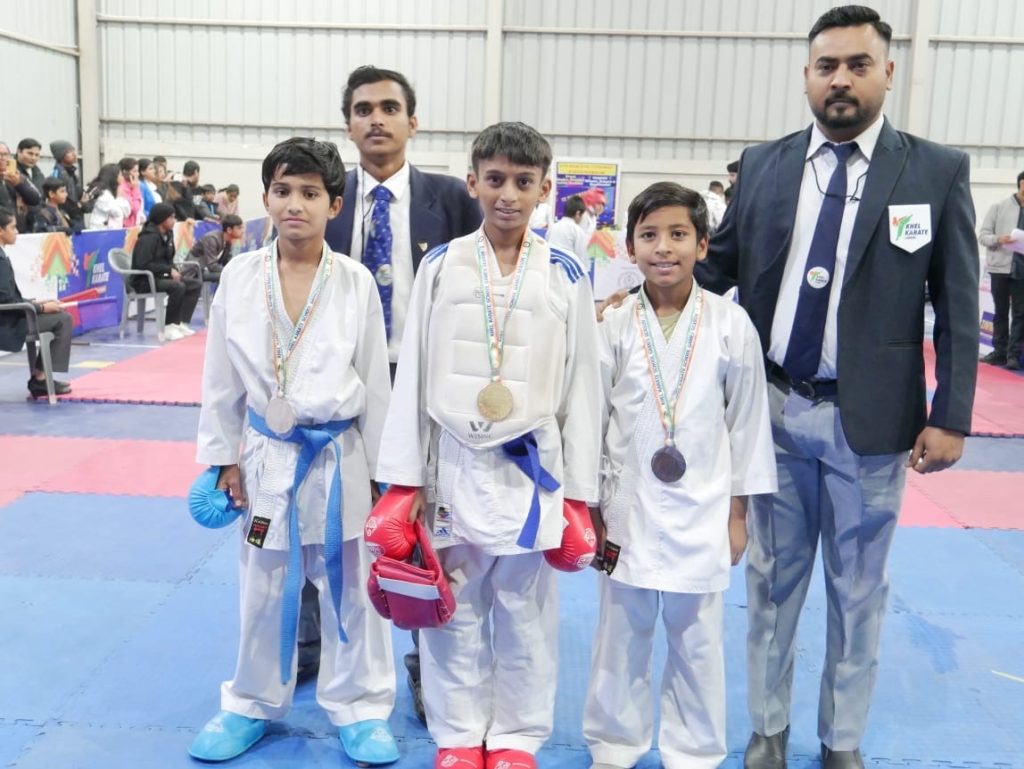 Three children wearing karate uniforms and medals stand on an indoor competition mat, accompanied by officials, during a national-level karate event.