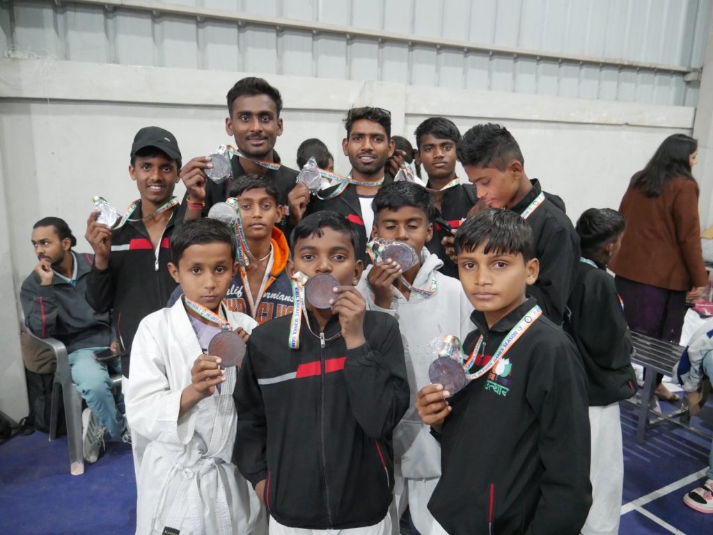 Group of children and youth from a martial arts training programme standing indoors and holding medals awarded at a national-level karate competition, with officials visible in the background.